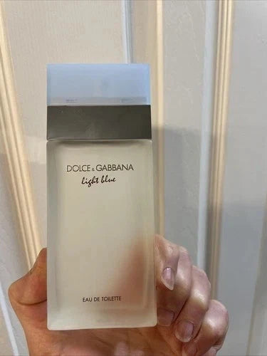 A hand holding a bottle of Dolce & Gabbana Light Blue Eau de Toilette against a white paneled door background.