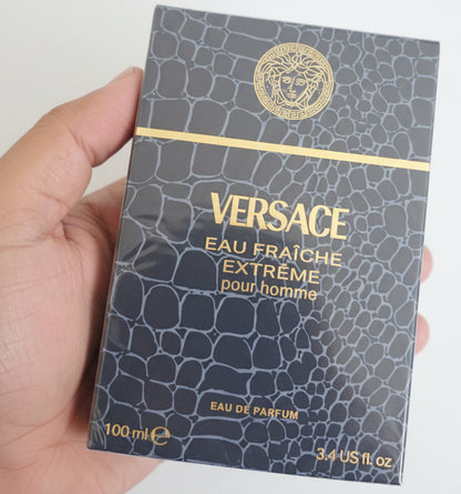 Versace Eau Fraçèche Extéreme pour homme perfume packaging held by a hand against a red background.