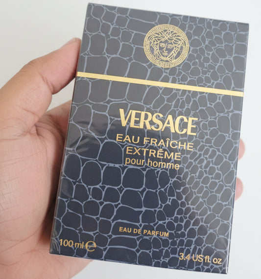 Versace Eau Fraçèche Extéreme pour homme perfume packaging held by a hand against a red background.
